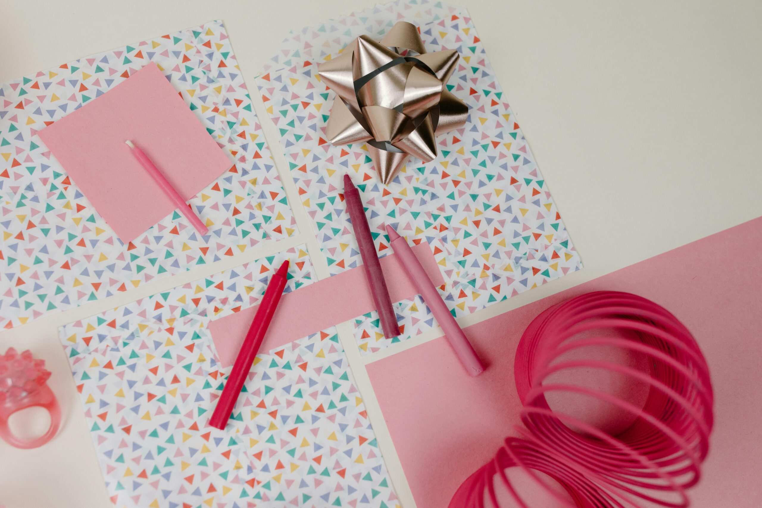 Top view of a vibrant creative arrangement featuring pink crayons, gift wrap, ribbon, and a slinky toy on a white background.