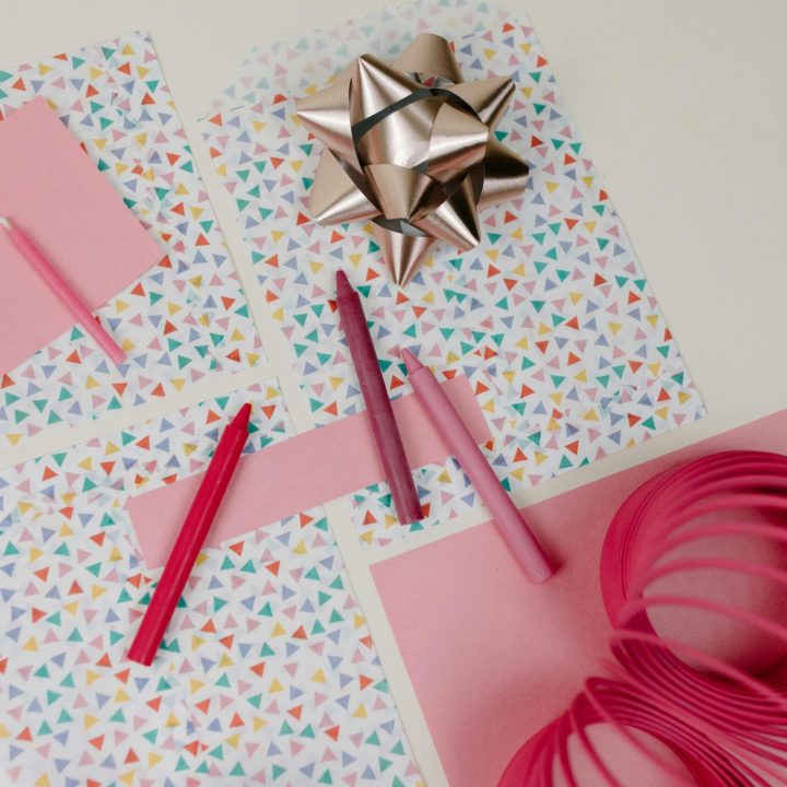 Top view of a vibrant creative arrangement featuring pink crayons, gift wrap, ribbon, and a slinky toy on a white background.
